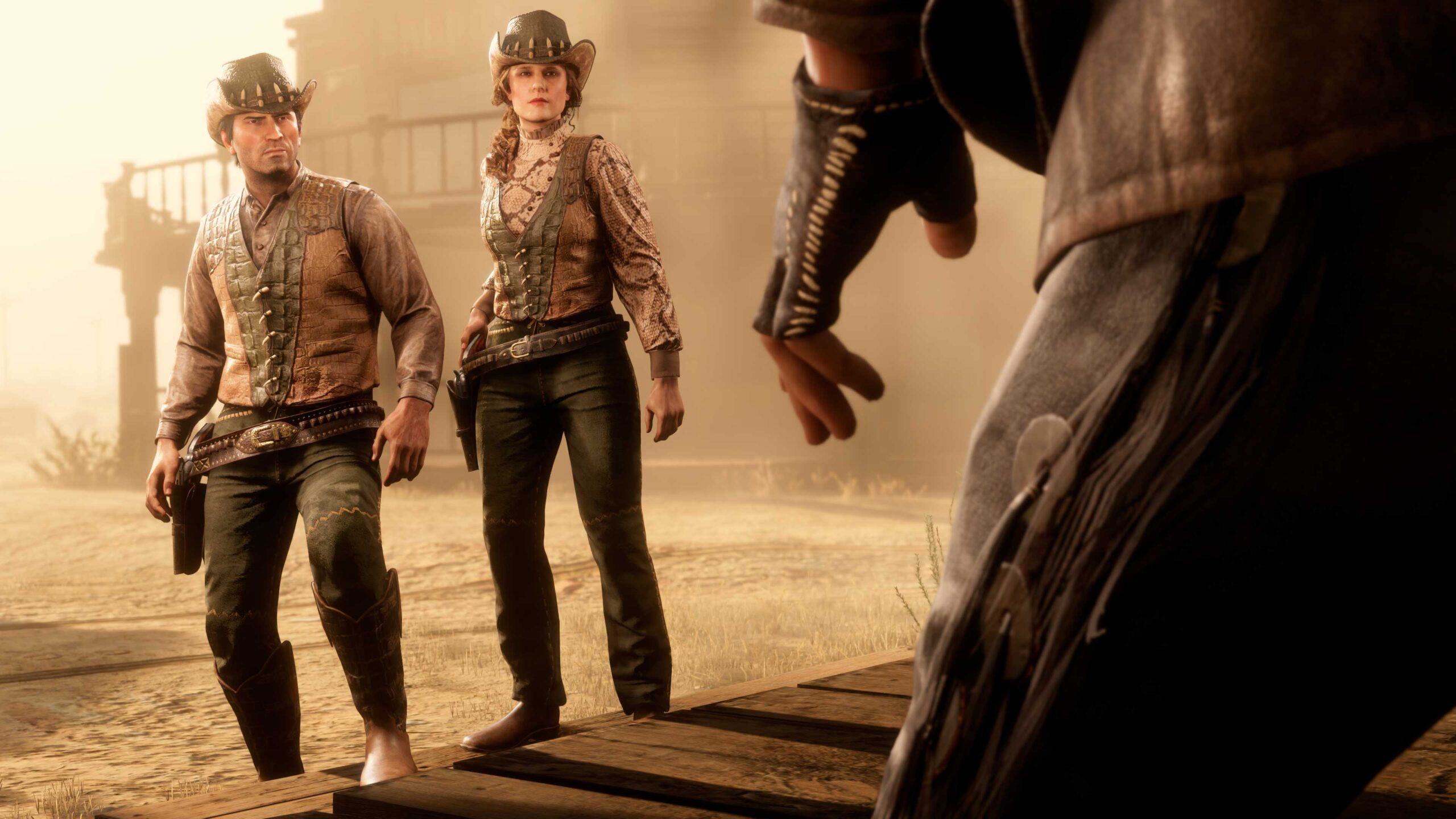 Um ângulo baixo mostra a mão do personagem próxima à cintura, pronta para puxar a arma. Dois outros personagens vestindo os Trajes inspirados na comunidade olham juntos para o primeiro personagem.